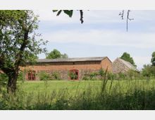 Eine ländliche Szene mit einem historischen roten Ziegelgebäude und einem danebenliegenden steinernen Wirtschaftsgebäude, umgeben von einer grünen Wiese und Bäumen. Die Architektur weist große, rote Tore und Fenster auf, typisch für eine Scheune oder ein landwirtschaftliches Anwesen.