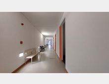 Ein langer, heller Flur mit weißen Wänden, einer hellen Decke und einem beigen Boden. Links stehen Holzbänke und rechts sind Türen, einige in Orange umrandet. Am Ende des Flurs ist eine Glastür sichtbar.