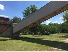 Eine industrielle Anlage mit einer schrägen, überdachten Brücke aus Wellblech, die über ein Gleis und eine Wiese verläuft. Im Hintergrund sind Bäume, ein Stück altes Backsteingebäude und Teile von technischen Maschinen zu sehen.