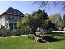 Ein grüner Garten mit einem Holztisch und Bänken im Vordergrund, daneben ein großer Baum. Im Hintergrund ein zweigeschossiges Haus mit Dachgauben und rotem Ziegeldach, umgeben von weiteren Bäumen und einer gepflegten Hecke.