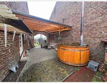 Innenhof zwischen zwei Gebäuden mit Wänden aus rotem Backstein, überdacht mit einer Holz- und Wellblechkonstruktion. Im Vordergrund ein runder holzverkleideter Whirlpool mit Kaminabzug, daneben Sitzmöbel und Deko-Elemente entlang der Hauswand.