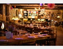 Rustikal eingerichteter Gastraum mit Holztischen und -stühlen, gedeckt mit Tellern und Gläsern. Im Hintergrund eine dekorative Bar mit bunten Lichtern, Wänden voller Bilder und einem Kronleuchter an der Decke.