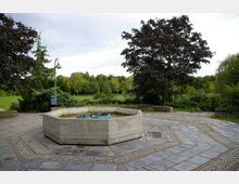 Ein leerer, achteckiger Betonbrunnen auf einem gepflasterten Platz, umgeben von Bäumen und Grünflächen in einem Park. Im Hintergrund sind dichte Bäume und Büsche zu sehen, und eine Laterne steht links neben dem Brunnen.