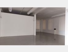 Leerer Innenraum mit weiß gestrichenen Wänden, einer glatten grauen Bodenfläche und sichtbaren Lüftungsrohren an der Decke. Der Raum hat große, gleichmäßig angeordnete Fenster mit teilweise durchscheinenden Vorhängen.