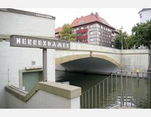Ansicht einer Brücke über einen Fluss in einer städtischen Umgebung. Im Vordergrund ist ein Schild mit der Aufschrift "Herrendamen" sowie ein Eingangsbereich zu öffentlichen Toiletten zu sehen; im Hintergrund befinden sich Wohngebäude mit roten Dächern und Bäumen.