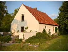Weißes, einstöckiges Haus mit rotem Satteldach in ländlicher Umgebung, umgeben von Bäumen und Garten. Vor dem Haus befinden sich eine Holzterrasse, Pflanzen und eine Sitzbank im Grünen.