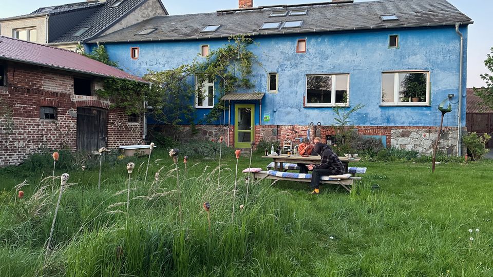 Hinterhof mit hohem Gras vor einem langgestreckten, blau verputzten Wohnhaus mit dunkel gedecktem Satteldach und mehreren Dachfenstern. Links steht ein niedriger Backstein-Nebengebäude mit Holztor, in der Mitte führt eine grüne Haustür ins Gebäude, daneben liegen eine Wiese und ein Picknicktisch.
