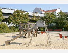 Spielplatz mit Sandboden, einer metallischen Rutsche, einem Klettergerüst aus Holz und roten Wipptieren. Im Hintergrund sind Bäume, eine Hochstraße sowie Gebäude mit roten und grauen Dächern zu sehen.