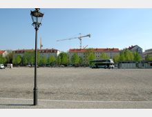 Gepflasterter Platz mit vereinzelten Personen und einem geparkten Reisebus; im Hintergrund befinden sich Wohnhäuser mit roten Ziegeldächern, Bäume, Baukräne und kleinere Pavillons. Im Vordergrund steht eine schwarze Laterne.