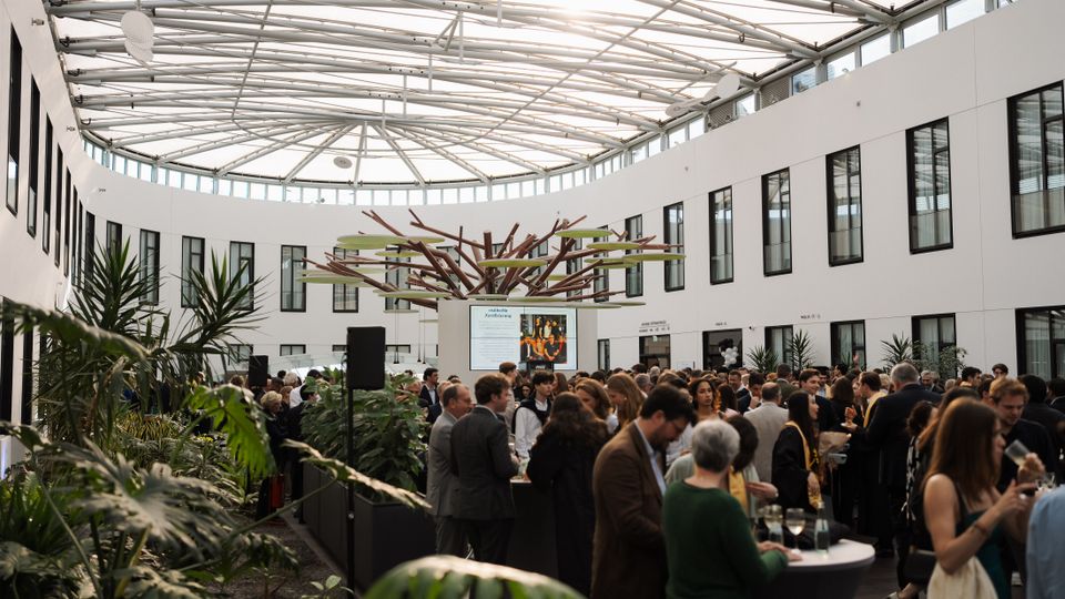 Geräumiger, moderner Veranstaltungsraum mit einer ovalen Glasdachkonstruktion, weißen Wänden mit rechteckigen Fenstern und einer zentralen Pflanzeninstallation in Baumform; am Rand des Raumes befinden sich Grünpflanzen und dazwischen sind Stehtische sichtbar.