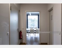 Flur in einem modernen Bürogebäude mit weißen Wänden, einer Tür beschriftet mit "WC" links, einem Feuerlöscher an der Wand und einer Glastür am Ende des Flurs, die in einen Besprechungsraum mit einem Tisch und Stühlen führt. Durch das Fenster im Hintergrund sind Bäume zu sehen.