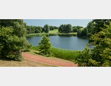 Ein ruhiger See in einer grünen Parklandschaft, umgeben von Bäumen und Schilf. Im Vordergrund verläuft ein gepflasterter Gehweg entlang des Ufers.