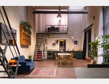 Wohnbereich im Industriestil mit offener Küche, hoher Decke und Zwischengeschoss. Sichtbare Metalltreppe, Holzboden, Pflanzen als Dekoration und modernes Mobiliar, darunter ein dunkelblaues Sofa, ein Esstisch mit gelben Stühlen und Küchenzeile.