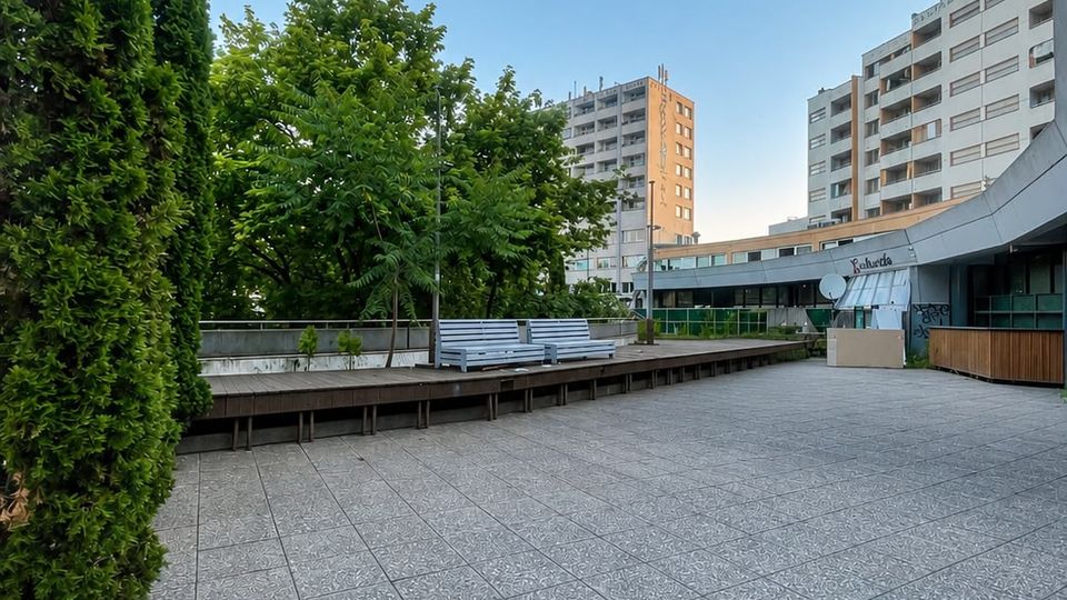 Großer, gepflasterter Außenplatz mit breiten Fliesen, einer erhöhten Holzplattform mit Sitzbänken und dichtem Baumbestand am Rand. Im Hintergrund stehen mehrere mehrstöckige Wohn- und Geschäftshäuser, rechts ein geschwungenes Gebäudedach mit Glasfront.