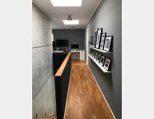 Ein moderner Flur mit Holzboden und grauen Wänden, an denen gerahmte Schwarz-Weiß-Bilder auf weißen Wandregalen hängen. Auf der linken Seite befindet sich eine Betonwand, während im Hintergrund Schreibtische mit Monitoren und Büromaterial erkennbar sind, unter einem von Oberlichtern beleuchteten Raum.