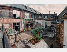 Ziegelstein-Innenhof eines alten Industriegebäudes mit renovierten Elementen und einer Mischung aus Beton-, Holz- und Fliesenböden. Der Hof ist mit Pflanzen, Holzstapeln und Sitzgelegenheiten dekoriert, und umliegende Gebäude haben große Fenster und metallische Balkongeländer.