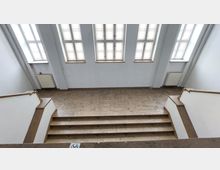 Treppenhaus mit breiten Steinstufen und weißen Wänden. Über den Stufen befinden sich mehrere große Fenster mit schlichtem Rahmen, die Tageslicht hereinlassen; auf beiden Seiten sind weiße Heizkörper angebracht.