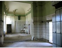 Ein leerer, heruntergekommener Raum mit hohen Decken, gefliesten Wänden und einem grünen Anstrich im oberen Bereich. Der Raum enthält Rohre, eine Türöffnung, ein Fenster, sowie einen Tisch und eine Spüle in der Ecke.