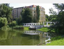 Ein kleiner Teich mit grünem Wasser, umgeben von dichtem Baumbestand und gepflegten Rasenflächen. Im Hintergrund ein mehrstöckiges gelbes Wohngebäude mit rotem Dach, daneben eine weiße Pergola mit einer kleinen Brücke über das Wasser.