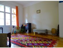 Helles Wohnzimmer mit Holzboden, einem großen bunten Teppich in geometrischem Muster, einem grauen Polsterhocker und einem schwarzen Regal an der Wand. Große Fenster mit orangefarbenem Vorhang lassen Tageslicht herein, daneben ein Blumentopf und ein Stativ.