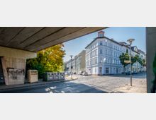 Straßenszene mit einem Eckgebäude im Gründerzeitstil, weiß gestrichen mit roten Dächern, umgeben von Kopfsteinpflasterstraßen. Im Vordergrund ist eine Brücke sichtbar, daneben stehen Müllcontainer und Bäume mit herbstlich verfärbtem Laub.