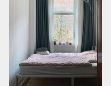 Ein schlichtes Schlafzimmer mit einem Bett in der Mitte, bedeckt von rosa Bettwäsche. Im Hintergrund ein großes Fenster mit Blick auf eine Backsteinwand und einige Zweige; das Fenster ist von dunkelgrünen und weißen Vorhängen umrahmt, und auf der Fensterbank stehen Blumentöpfe sowie kleine Gegenstände.