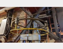 Industrieller Innenraum mit sichtbarer Ziegelwand, Stahlträgern und Seilen. Im Vordergrund eine Maschine mit einem gelben, kreisförmigen Metallring und mechanischen Bauteilen.