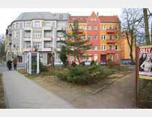 Straßenszene mit mehrstöckigen Wohnhäusern im Hintergrund, die in grau-weißen und orange-roten Farben gestrichen sind. Im Vordergrund befindet sich eine kleine Grünfläche mit niedrigen Büschen, ein Gehweg, eine Telefonzelle und ein Plakat an einem Zaun.