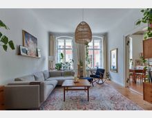 Wohnzimmer mit einem großen grauen Ecksofa, einem Holzcouchtisch und einem gemusterten Teppich. Drei hohe Fenster lassen Tageslicht hinein, dekoriert mit Pflanzen und hellen Vorhängen, ergänzt von einer hängenden Korblampe sowie einem angrenzenden Essbereich im Hintergrund.