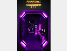 Raum mit futuristischem Thema, beleuchtet von violetten Neonlichtern, dargestellt durch einen hexagonalen Eingang mit der Aufschrift „Gateway Station – Authorized Personnel Only“. Im Inneren befinden sich Computerterminals und Bildschirme in einer sci-fi-ähnlichen Kontrollraum-Ästhetik.