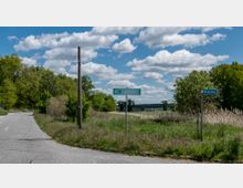 Ländliche Straßenszene mit einer asphaltierten Straße, die nach links abbiegt, umgeben von grünen Feldern und Bäumen. Straßenschilder zeigen "Blumenweg" und eine allgemeine Richtung, während im Hintergrund ein bewölkter blauer Himmel zu sehen ist.