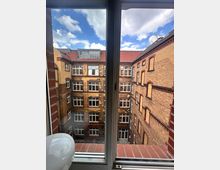 Blick aus einem Fenster auf den Innenhof eines historischen Backsteingebäudes mit mehreren Stockwerken und weißen Sprossenfenstern. Der Himmel ist blau mit einigen weißen Wolken.