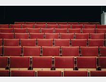 Reihen von roten, gepolsterten Sitzen in einem leeren Theater- oder Kinosaal vor einem dunklen Hintergrund.