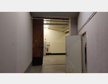 Flur mit schlichten weißen Wänden und einem offenen Durchgang, der zu einem weiteren Raum mit gelblicher Wand führt. Im Hintergrund sind Feuerlöscher sowie eine Beleuchtung mit sichtbaren Rohren an der Decke erkennbar.