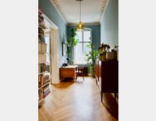 Ein heller Raum mit Fischgrätenparkett, einer hohen Decke mit Stuckverzierungen und einer Hängelampe. Im Hintergrund steht ein Schreibtisch vor einem großen Fenster, umgeben von zahlreichen Zimmerpflanzen; links Bücherregale, rechts ein Holzschrank.