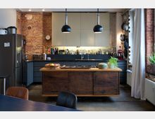 Moderne Küche mit einer Kombination aus unverputzter Ziegelwand und schwarzen Schrankfronten. In der Mitte steht eine rustikale Kücheninsel aus Holz, umgeben von Hängelampen, während Regale gefüllt mit Gläsern und Kochutensilien sichtbar sind.