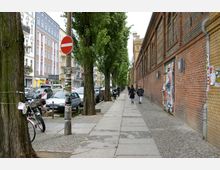 Straßenszene in einem urbanen Wohngebiet mit einer gepflasterten Gehwegfläche zwischen einer Reihe von Bäumen und einer Backsteinmauer rechts, die mit Plakaten bedeckt ist; links stehen parkende Autos vor mehrstöckigen Gebäuden. Im Hintergrund spazieren zwei Personen entlang des Gehwegs.