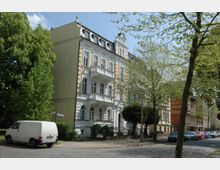 Mehrstöckiges Altbaugebäude mit heller Putzfassade und dekorativen Balkonen in einer von Bäumen gesäumten Stadtstraße. Im Vordergrund steht ein weißer Lieferwagen auf Kopfsteinpflaster, daneben geparkte Autos entlang der Straße.