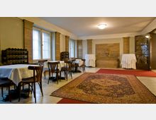 Innenraum eines historisch gestalteten Raumes mit Holzmöbeln und weiß gedeckten Tischen. Der Raum hat beige Wände, dekorative Säulen, große Fenster mit Vorhängen und einen zentralen Teppich auf dem Boden.