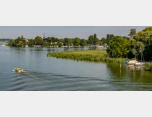 Flusslandschaft mit bewaldeten Ufern, Segelbooten am rechten Bildrand und einer kleinen Insel mit Gebäuden und einer Kirche im Hintergrund; ein Motorboot fährt links durch das Wasser.