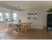 Esszimmer mit einem großen Holztisch und acht passenden Holzstühlen auf hellem Holzboden. An der grauen Wand hängen sechs schwarz-weiße Bilder, links befinden sich drei Fenster mit Pflanzen und einer Tischleuchte. Ein moderner Kronleuchter hängt über dem Tisch, rechts steht ein schwarzer Schrank und ein Korb.