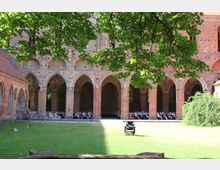 Historische Klosteranlage mit roten Backsteinbögen und gotischen Arkaden, im Hintergrund sitzen Menschen auf Bänken unter den Arkaden, im Vordergrund eine grüne Rasenfläche mit Baumzweigen.