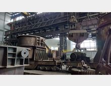 Eine große Industriehalle mit schweren Maschinen und Stahlkonstruktionen, darunter ein massiver Schmelztiegel auf Rollen und verschiedene Kräne. Im Hintergrund sind große Fenster und Metallträgerstrukturen sichtbar.