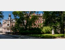 Historisches Gebäude mit verzierten Fassaden, Türmen und Zierelementen, teils von Bäumen und Sträuchern in einem parkähnlichen Umfeld verdeckt; im Vordergrund eine Büste auf einem Steinsockel.