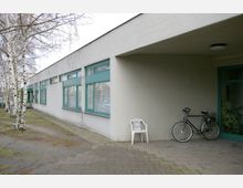 Flacher, moderner Gebäudekomplex mit cremefarbener Fassade und großen, grünen Fensterrahmen. Im Vordergrund stehen eine weiße Plastikstühle und ein Fahrrad unter einem überdachten Bereich, daneben Birkenbäume und gepflasterte Wege.