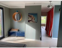 Heller Innenraum eines modernen Hauses mit blassgrünen Wänden, einem runden Wandspiegel mit Holzrahmen und einem blauen Schrank mit Sitzkissen davor. Im Hintergrund sind ein roter Vorhang, ein Fenster mit Blick ins Grüne und helle Holzböden zu sehen.