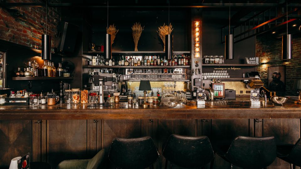 Innenansicht einer Bar im Industrial-Stil mit langer Holztheke, Barhockern und Regalen voller Spirituosen- und Glasflaschen. Sichtmauerwerk, dunkle Decke mit hängenden zylindrischen Lampen und warmes Akzentlicht prägen den Raum.