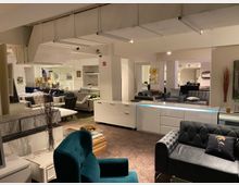 Innenansicht eines modernen Möbelgeschäfts mit verschiedenen Polstermöbeln wie Sofas und Sesseln, die in beleuchteten Ausstellungsräumen präsentiert werden. Der Raum ist mit weißen Wänden, Teppichboden und einer warmen Beleuchtung gestaltet, ergänzt durch dekorative Accessoires.