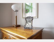 Holzkommode mit glänzender Oberfläche, darauf eine dekorative Tischlampe mit Holz- und Metallfuß sowie ein Fotorahmen mit verziertem Silberrand und einem schwarz-weißen Foto. Im Hintergrund hängt ein gerahmtes, abstraktes Bild an der weißen Wand.