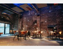 Industrieller Innenraum mit unverputzten Backsteinwänden, mehreren Tischen und Holzstühlen, die gleichmäßig im Raum verteilt sind. Große Fenster auf der linken Seite lassen Tageslicht herein, während einzelne Hängelampen die Tische beleuchten und eine gemütliche Atmosphäre schaffen.